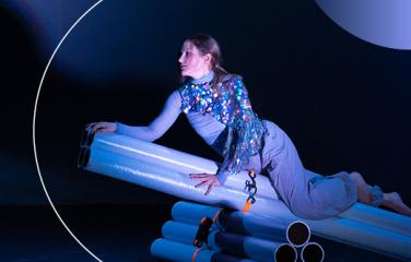 A performing artist dressed in purple clothing rests on a prop made of piping, and is illuminated by a spotlight.