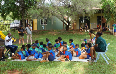 School children sit on the grass in front of a teacher during a language class. 