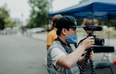 A screen worker on set holding a video camera with microphone and tripod. They are wearing a baseball cap and facemask.